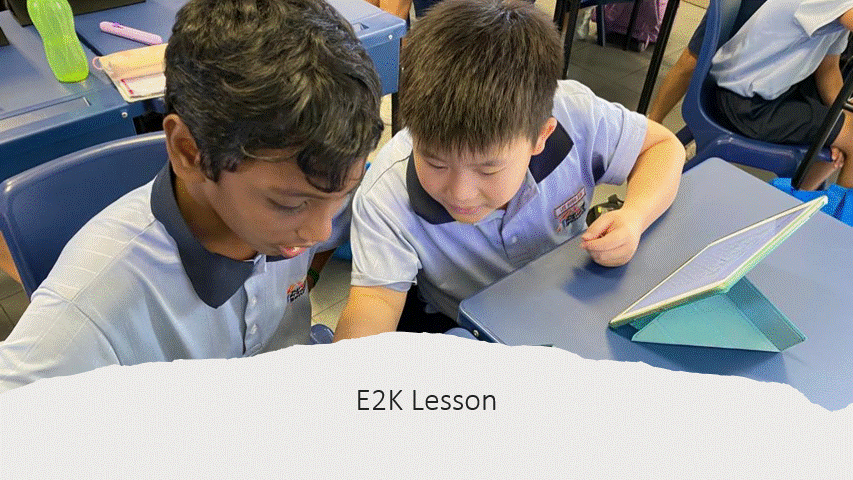 Two school boys in uniforms look at a tablet together at a classroom desk during an E2K lesson.